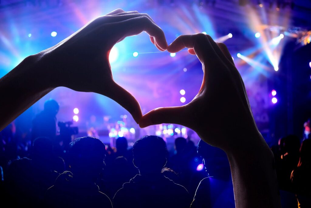 Hands forming a heart shape at a lively concert with colorful lights.